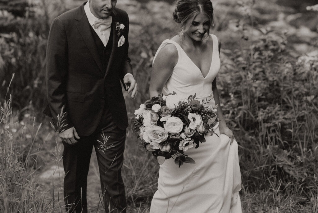 Photo of bride and groom walking through tall grass and bride is holding a bouquet